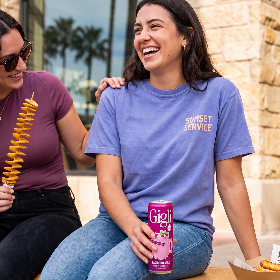 Two friends laughing at an outdoor table with a Raspberry Mule SKINNY Mocktail can